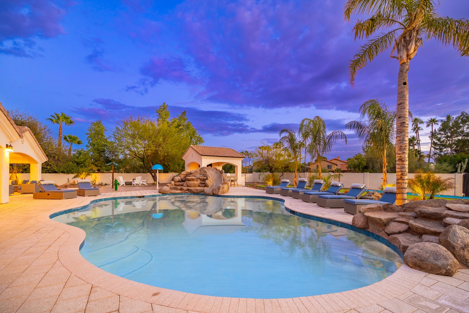 A stunning pool area surrounded by swaying palms and lounge chairs awaits, as twilight casts its colorful glow over this desert oasis paradise.