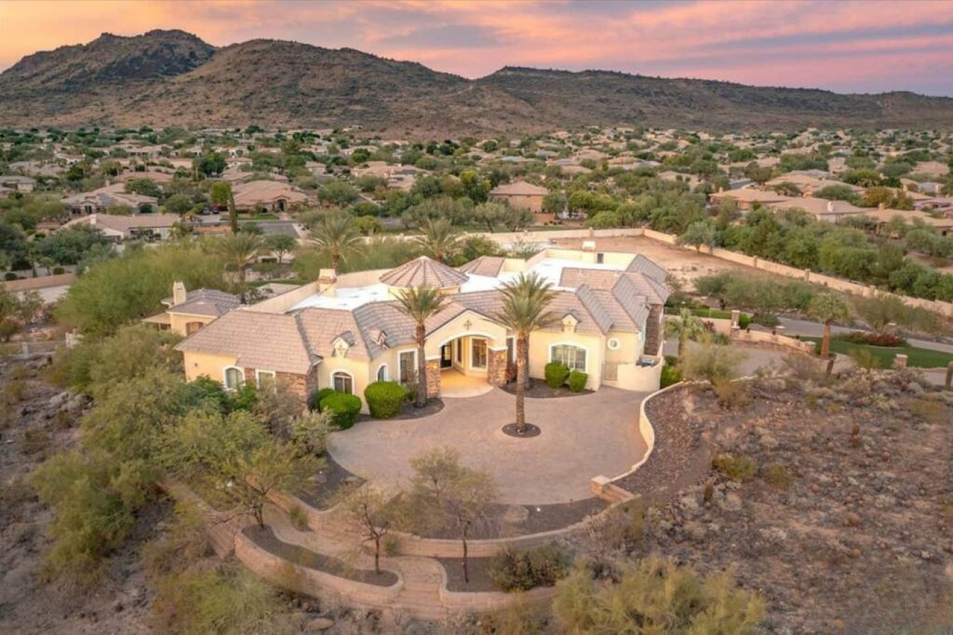 Aerial view of a spacious desert estate with mountain backdrop at sunset, surrounded by natural desert landscape and quiet residential neighborhood.