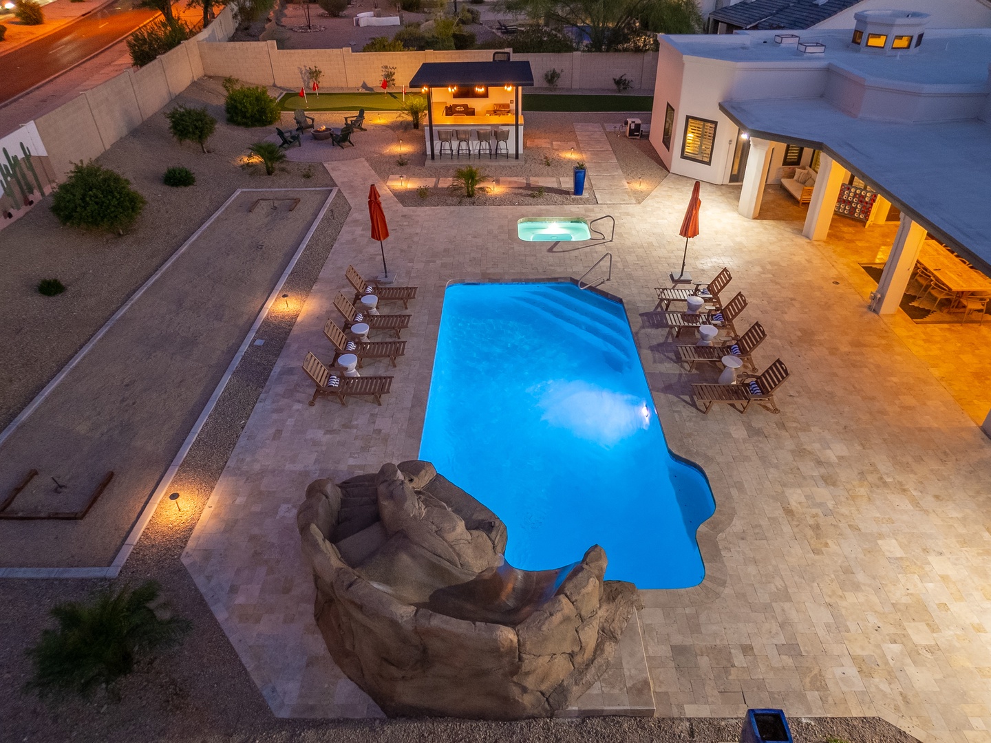Aerial view of the property showcasing a striking swimming pool with unique rock feature, hot tub, outdoor dining pavilion, and desert landscaping illuminated at dusk.