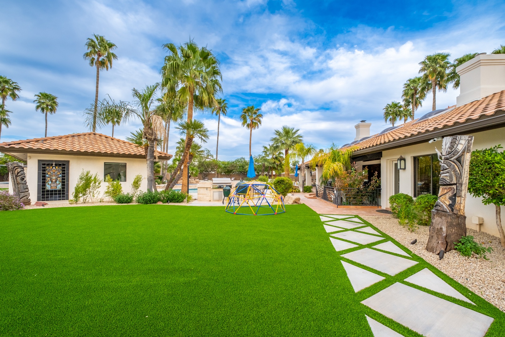 Spacious vacation home features vibrant landscaping with towering palms, a pristine lawn, climbing dome for kids, and inviting outdoor living areas perfect for sunny getaways.