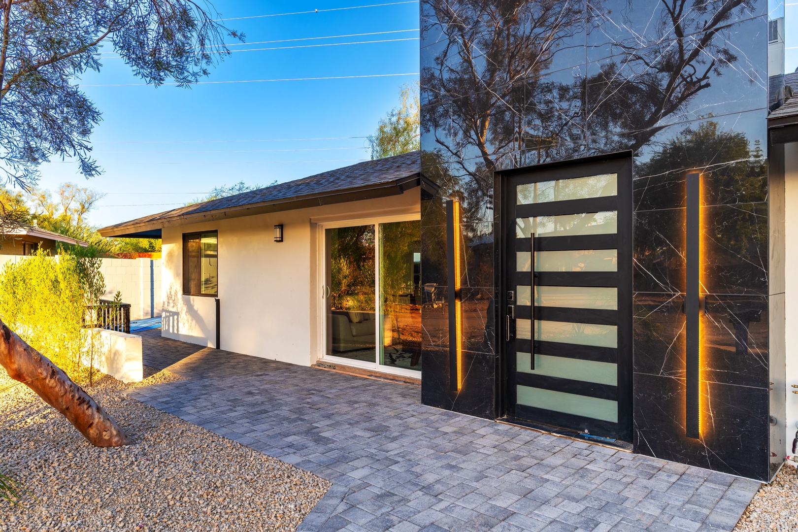 Modern property entrance featuring sleek black gates with decorative lighting, leading to a contemporary exterior framed by mature desert landscaping and clear blue skies.