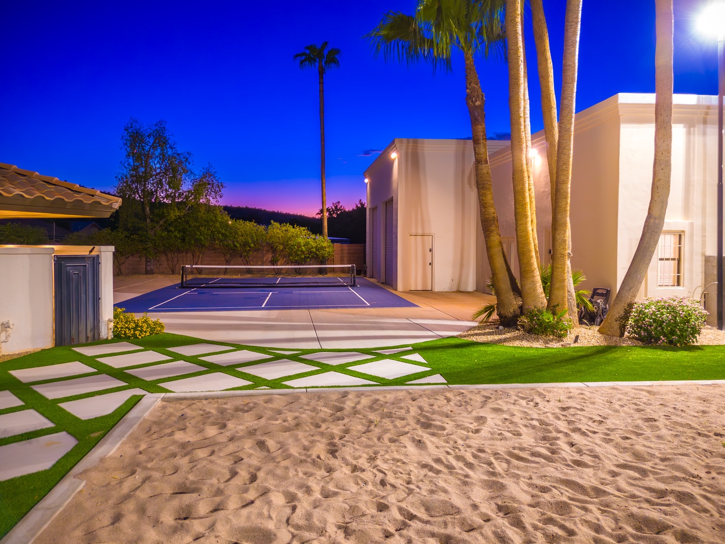 Private tennis court with illuminated play area, sand volleyball pit, lush palm trees, and modern architectural details under a twilight sky.