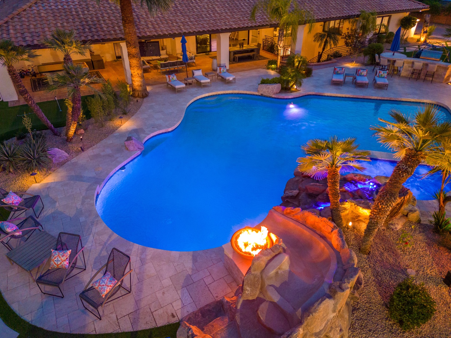 A stunning resort-style pool glows under the evening sky, complete with a fire feature, waterfall grotto, palm-lined deck, and multiple lounging areas—your perfect backyard oasis awaits.