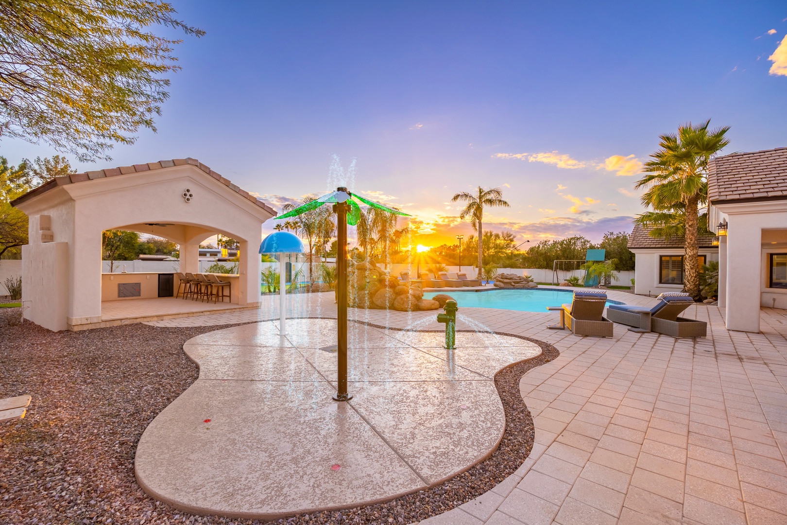 Sunlit backyard oasis with splash pad, pool, and covered patio—perfect for family fun and desert relaxation under swaying palms.