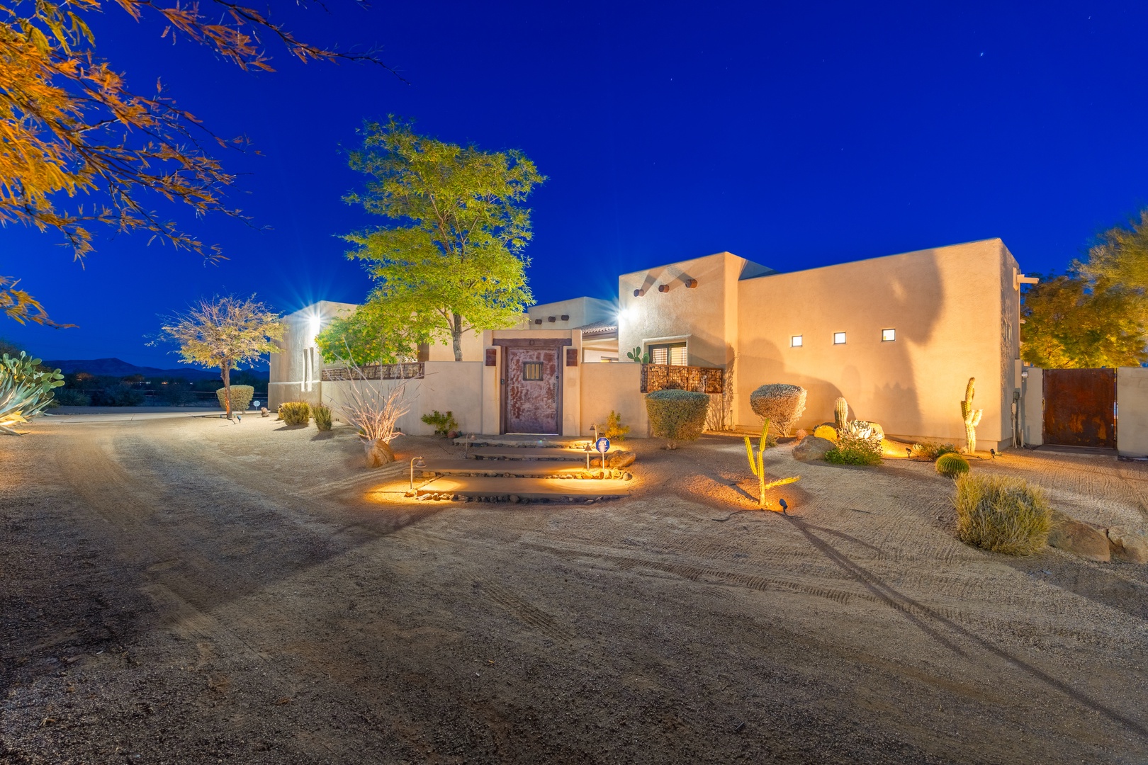Southwestern adobe architecture set against a twilight desert sky, surrounded by native landscaping and illuminated pathways leading to the carved wooden entrance.