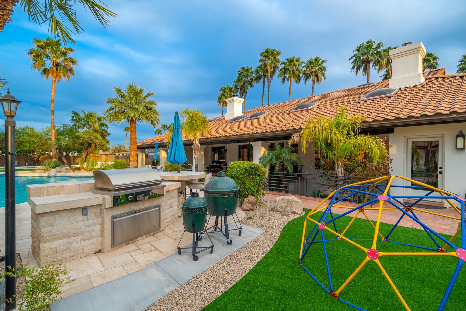 A resort-style paradise awaits with a sparkling pool, outdoor kitchen, BBQ area, and vibrant playground—all framed by swaying palms and mountain views, perfect for family getaways.