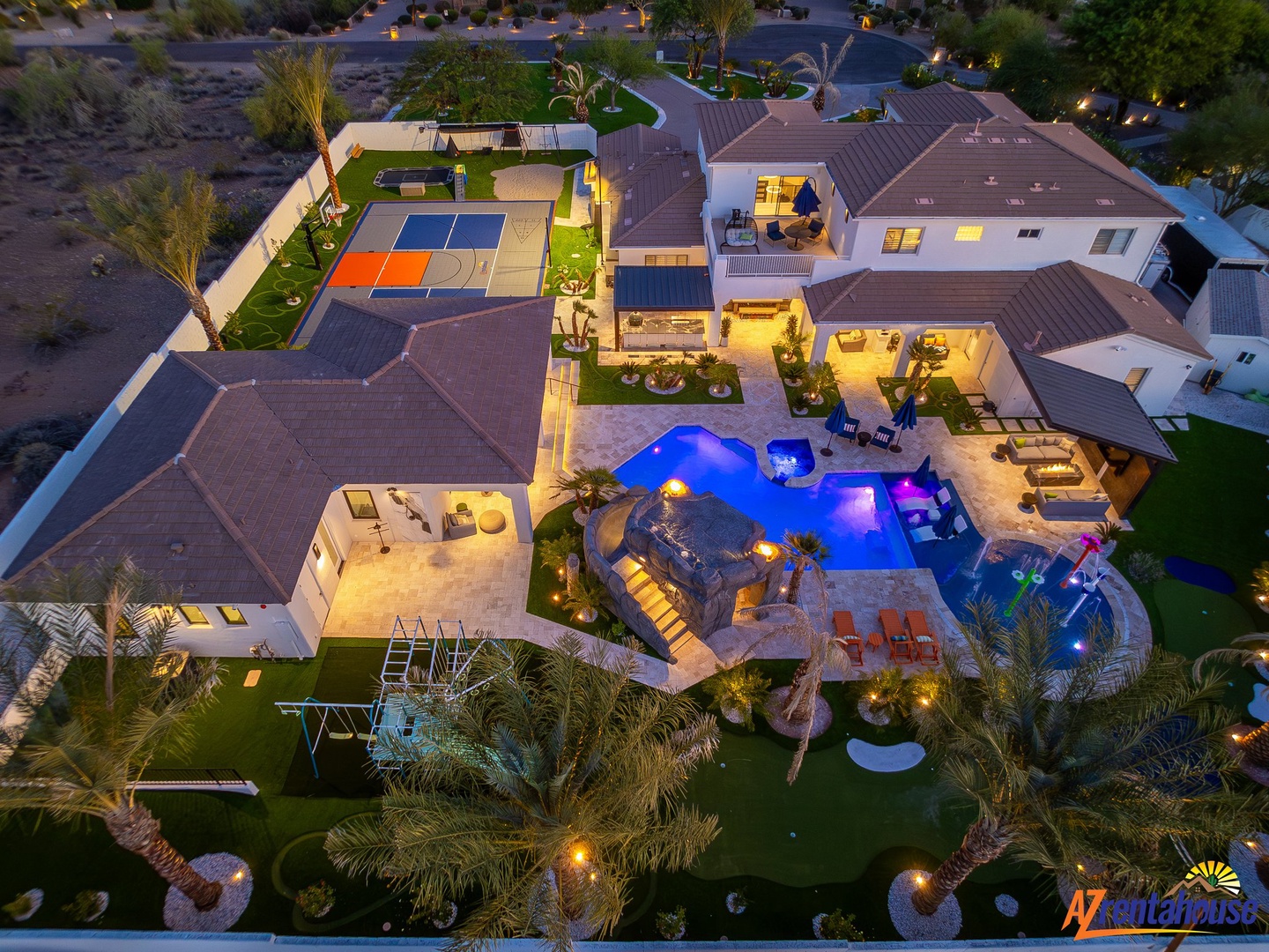 Stunning aerial view of a luxurious desert oasis featuring an illuminated pool and sport court in a beautifully landscaped setting.