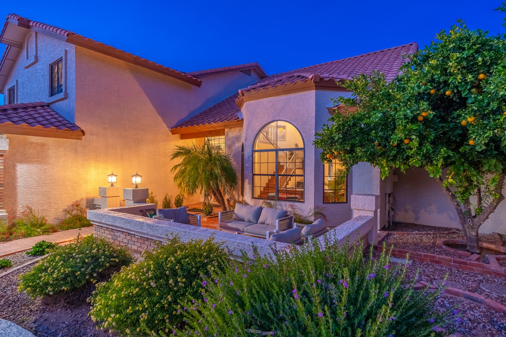 A charming stucco villa with Spanish tile roofing and an inviting courtyard patio. Warm evening lighting highlights the arched windows, landscaped gardens with citrus trees, and comfortable outdoor seating areas perfect for relaxation.