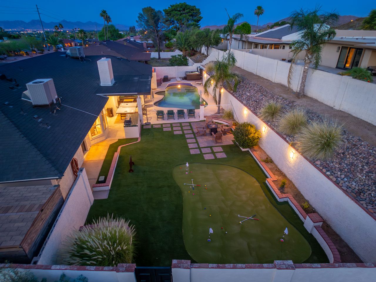 Aerial view of a resort-style backyard featuring a private putting green, heated pool, and covered patio in a quiet residential neighborhood surrounded by desert landscaping and mountain views.