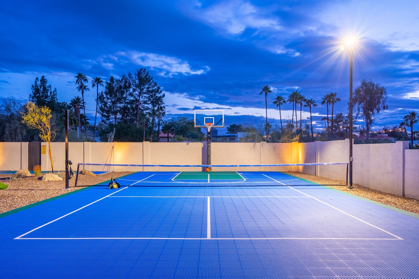Multi-purpose sport court featuring tennis and basketball under dramatic twilight skies, surrounded by tall palms and soft lighting for evening play.
