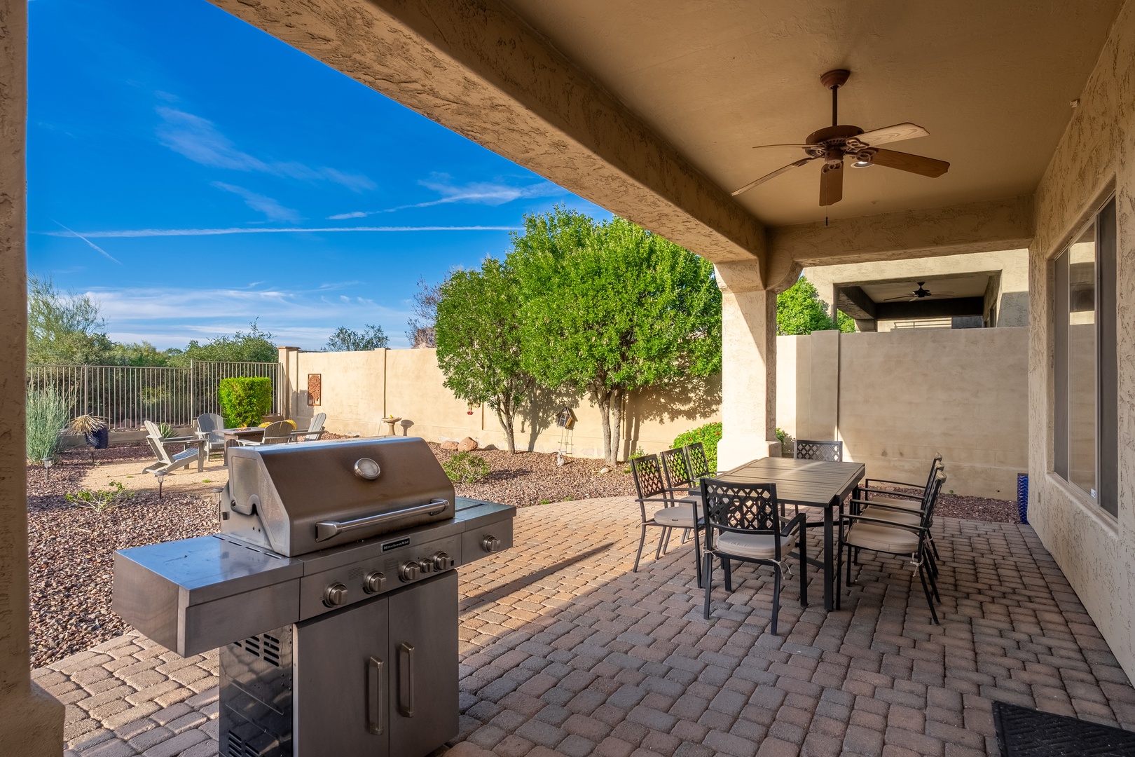A covered patio with a gas grill, dining table for six, ceiling fan, and views of the landscaped backyard with desert plants and clear blue skies above.