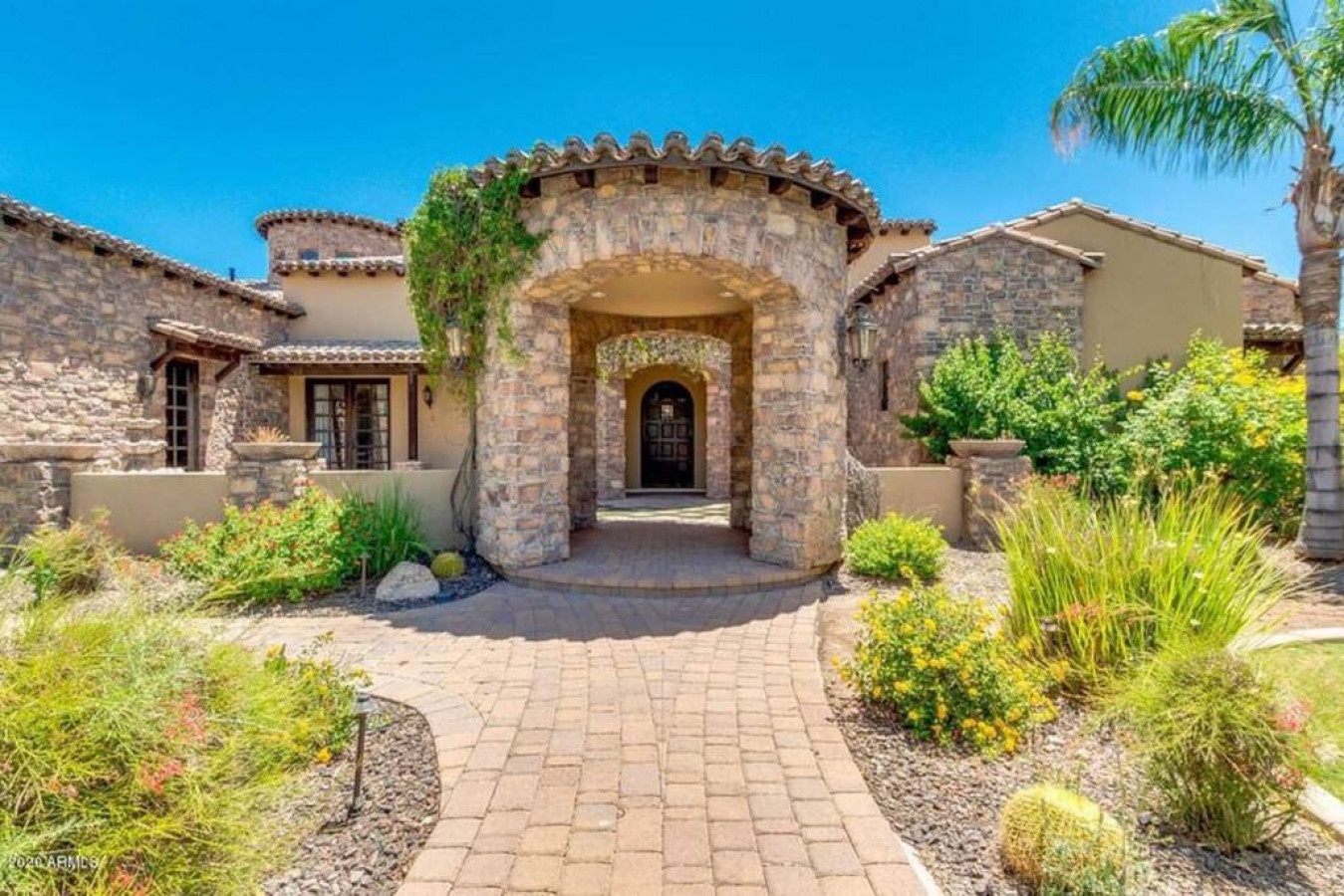 Stunning stone facade welcomes guests with Mediterranean charm, featuring beautiful desert landscaping and elegant archway entrance.