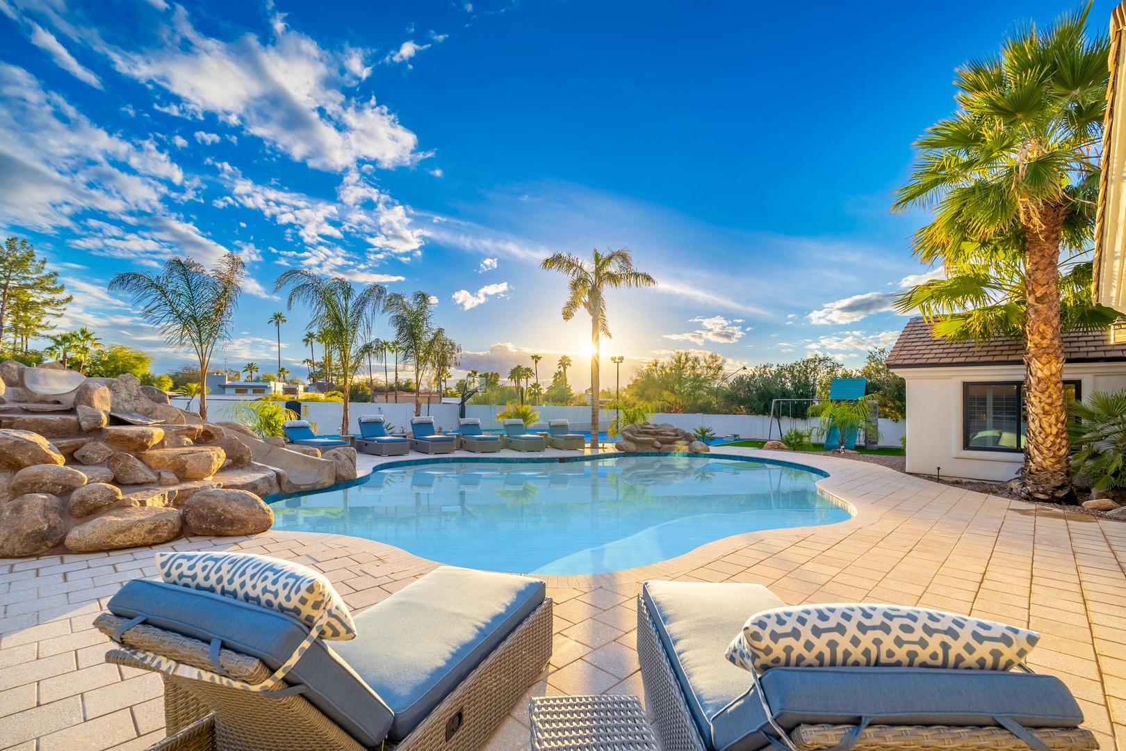 Expansive resort-style pool with cascading rock waterfall, palm trees, and plush lounge chairs set beneath golden sunlight and a sprawling blue sky.