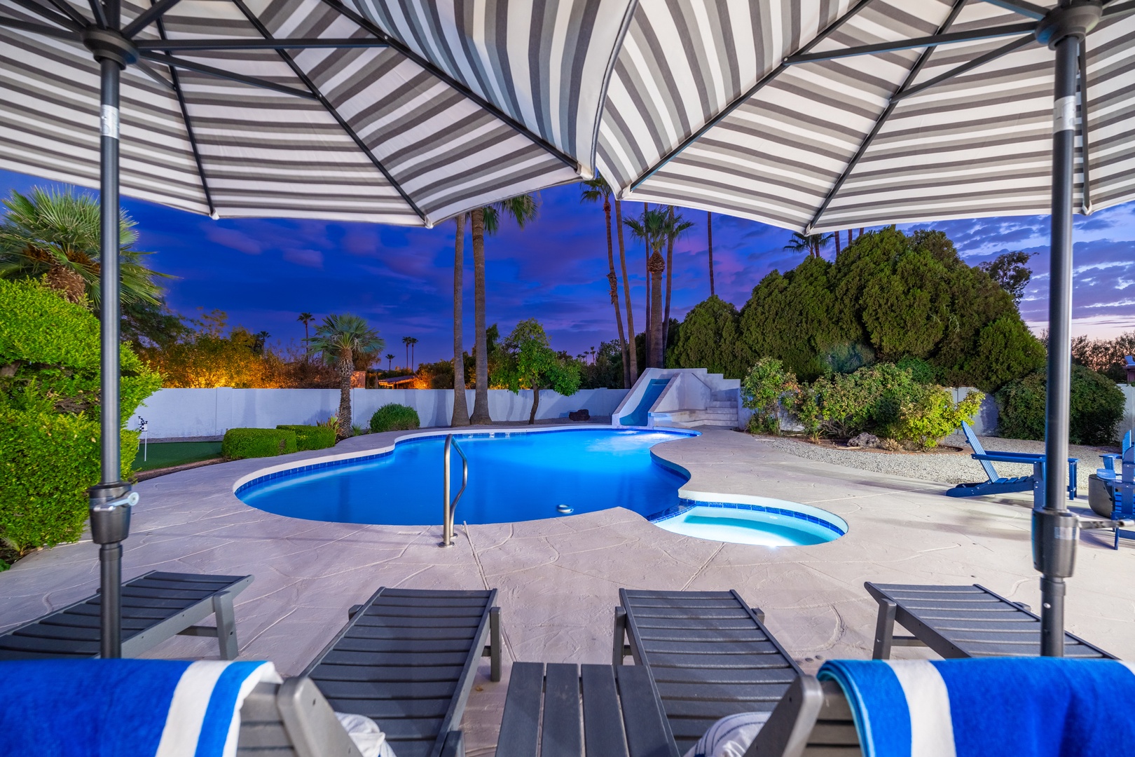 Relax under the stylish striped canopy as the pool glows against the twilight sky, surrounded by lush palms and manicured greenery.