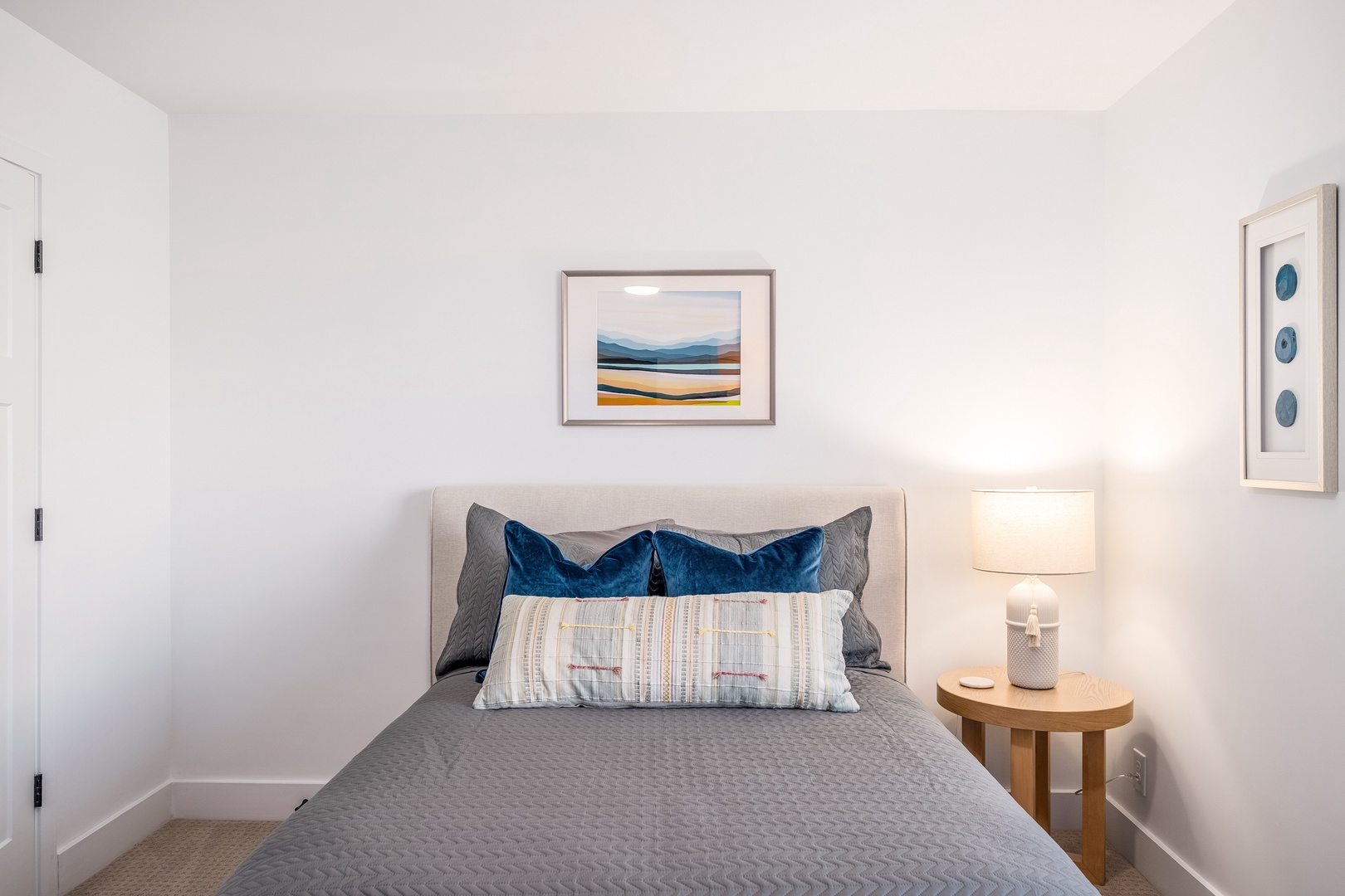 Your serene bedroom features plush bedding with velvet blue accent pillows, warm bedside lighting, and soothing coastal artwork—perfectly designed for restful nights.