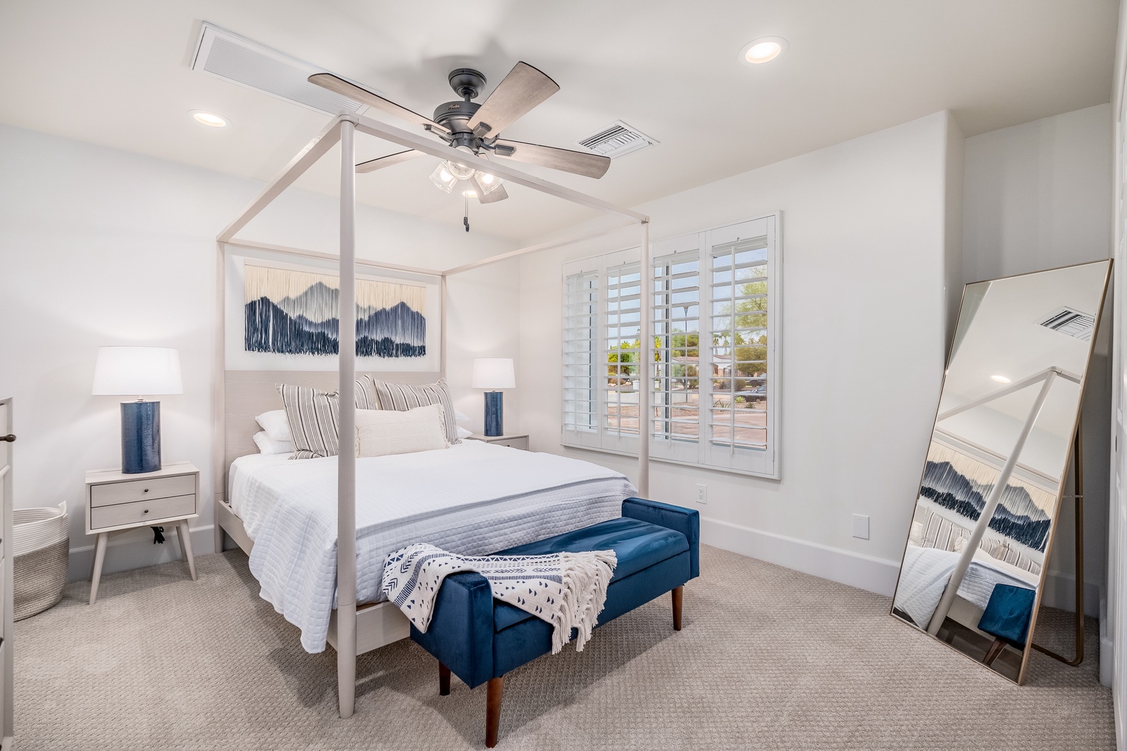 Your serene bedroom retreat features a plush canopy bed dressed in crisp white linens, complete with a cozy bench for unwinding. Soft natural light filters through plantation shutters while a ceiling fan keeps you comfortable.