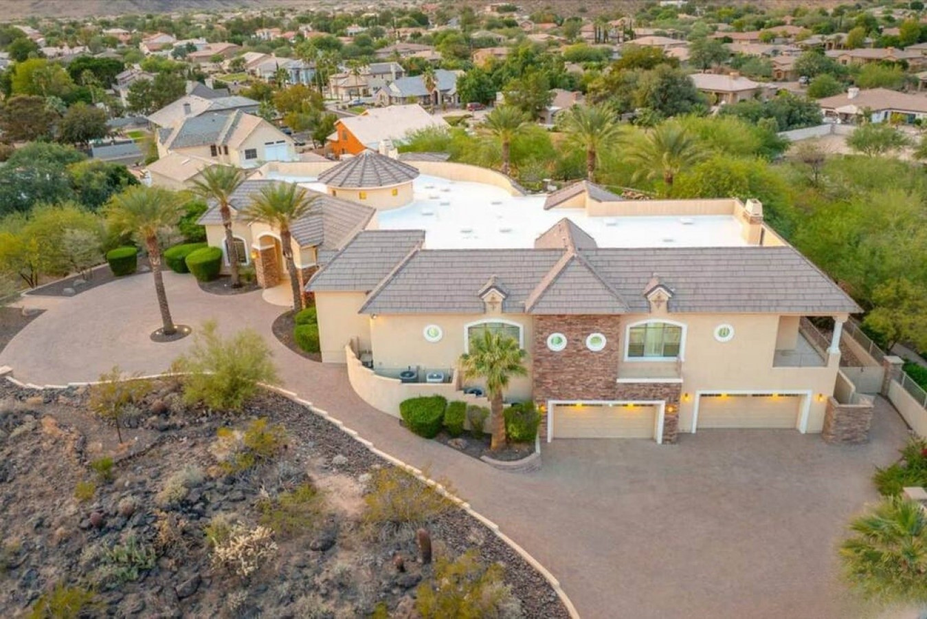 Aerial view of a spacious desert retreat featuring a circular driveway, palm-lined landscaping, and multi-level architecture set within a peaceful residential neighborhood.