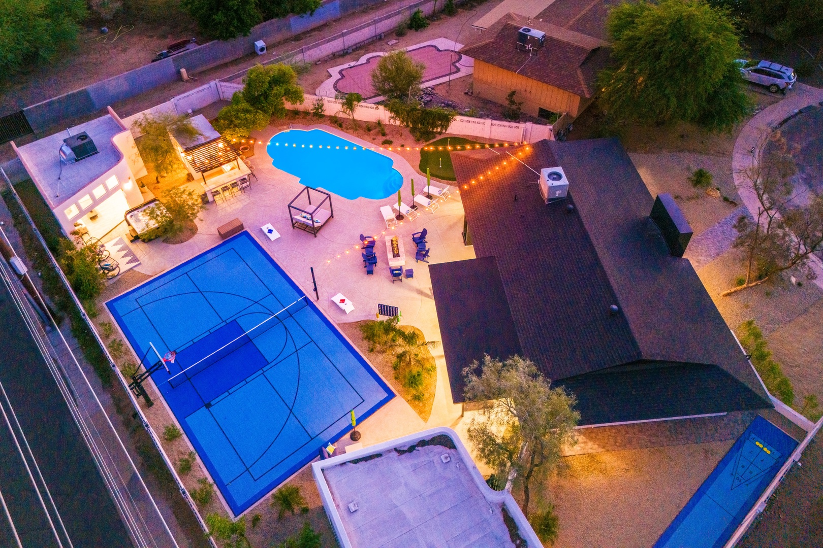 Aerial view of a vacation property featuring a swimming pool, sport court, and outdoor entertainment area illuminated by evening lighting, set in a serene desert landscape.