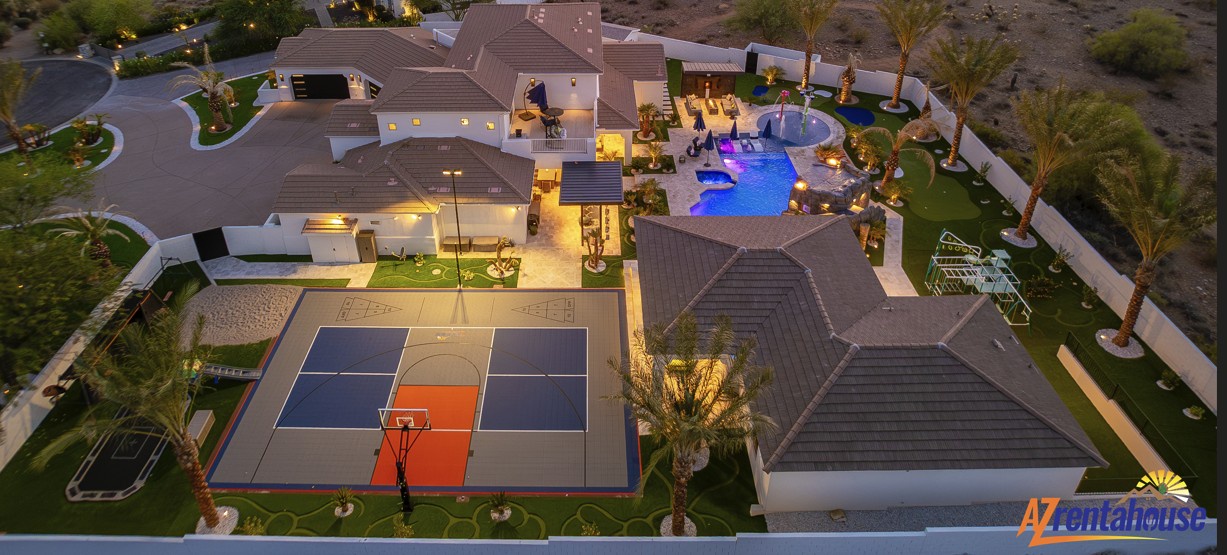 Luxurious estate sprawls across desert landscape with resort-style pool, full basketball court, and manicured gardens illuminated at dusk.