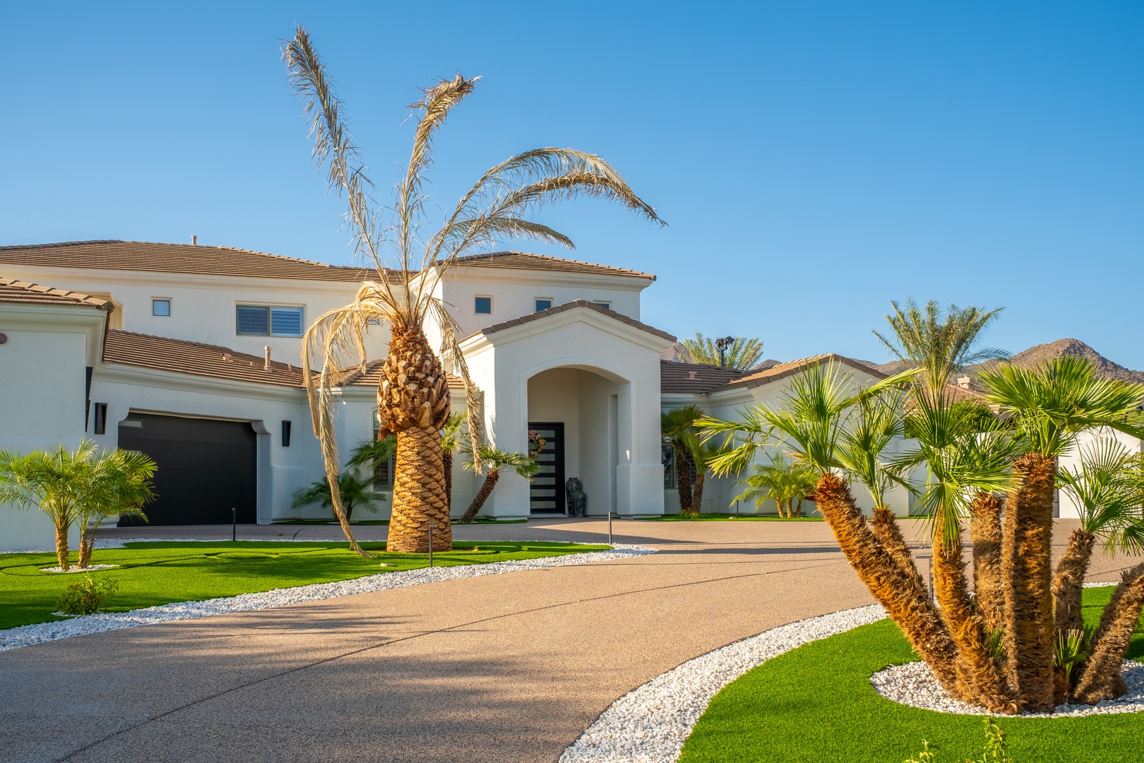 A modern desert retreat with elegant arched entryway, framed by mature palms and manicured landscaping under clear blue skies—your welcoming arrival awaits.