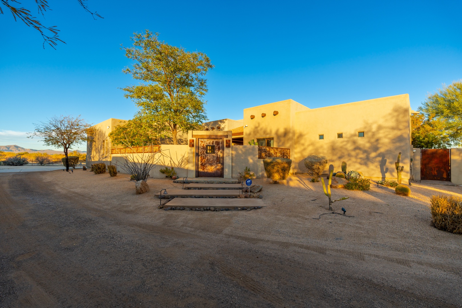 Adobe-style architecture bathed in golden light amid desert landscaping, native trees, and distant mountain views at sunset.