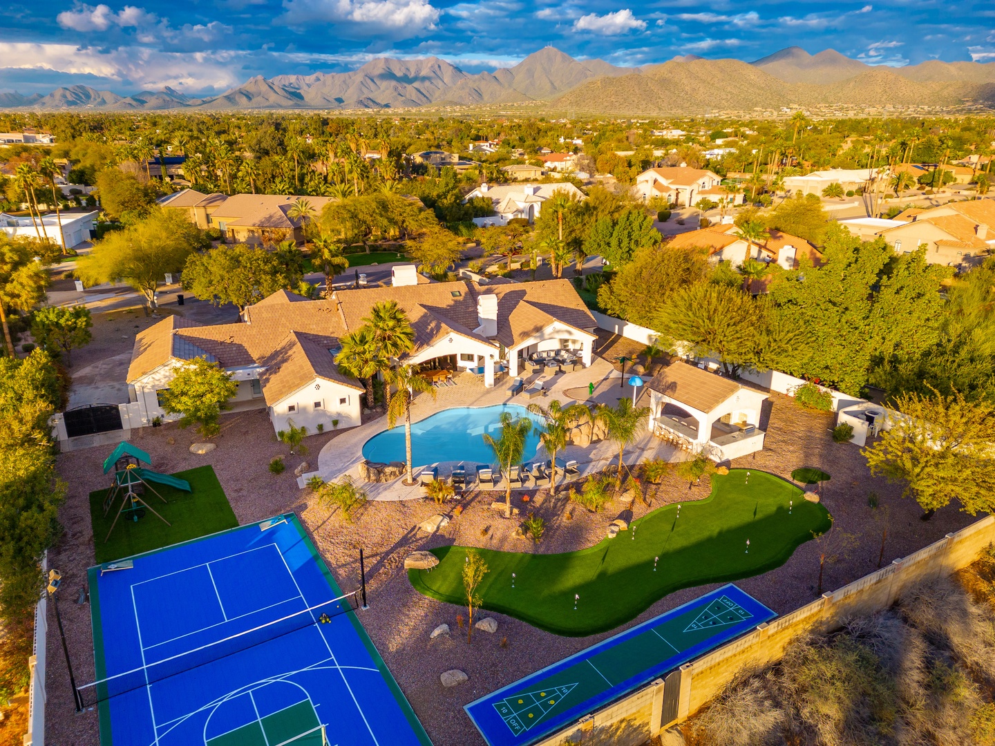 Aerial view of a desert vacation estate featuring a pool, private tennis and sport courts, mini golf, and spacious outdoor entertaining areas set against majestic mountain scenery.
