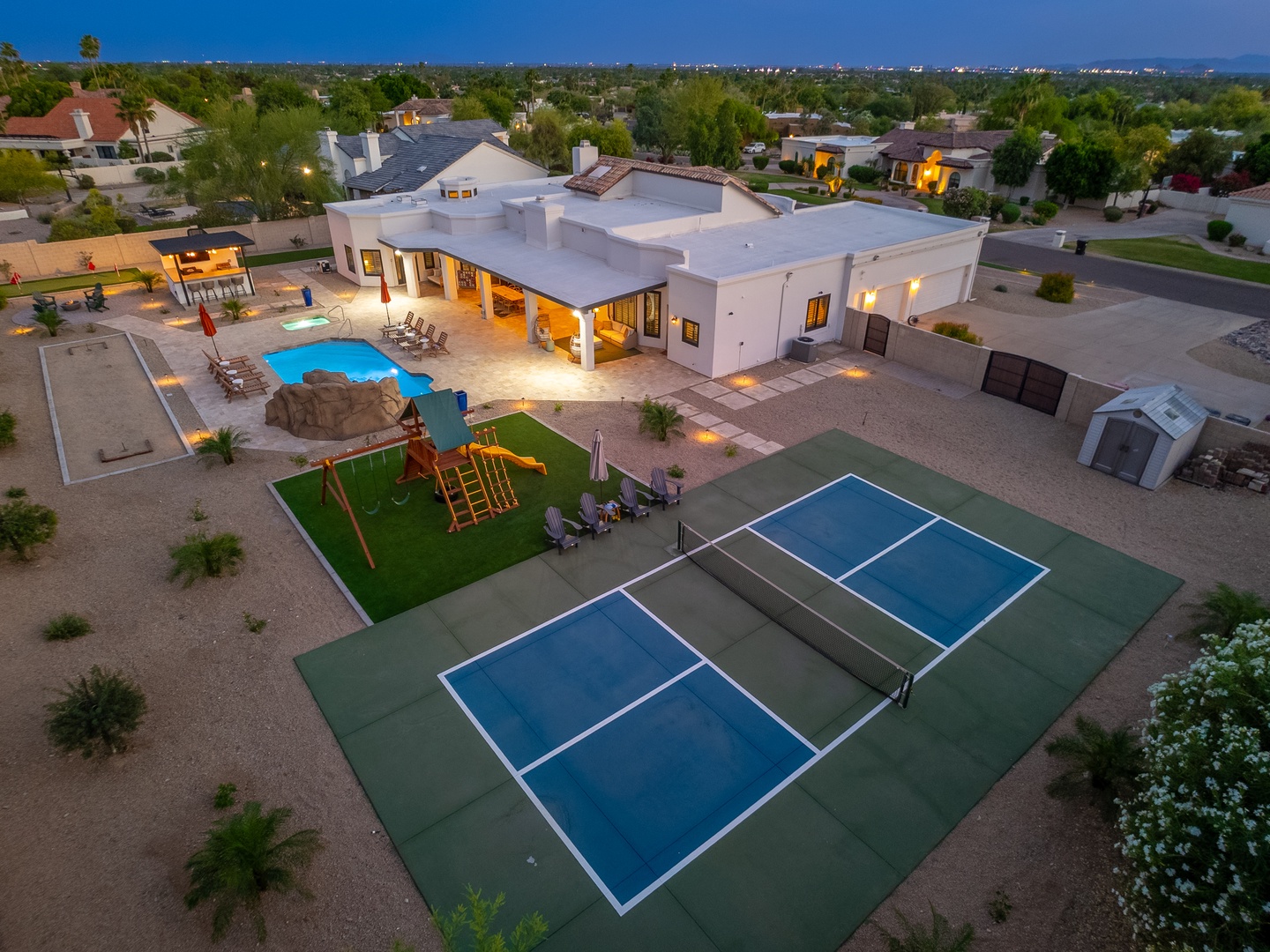 Aerial view of an expansive vacation property featuring a swimming pool, tennis court, playground, and covered patio area in a spacious residential neighborhood at twilight.