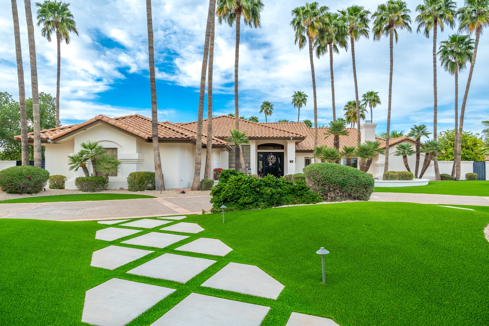 A stunning ranch-style home framed by towering palms and manicured landscaping, offering a private desert oasis with lush greenery and a welcoming entry in a serene residential neighborhood.