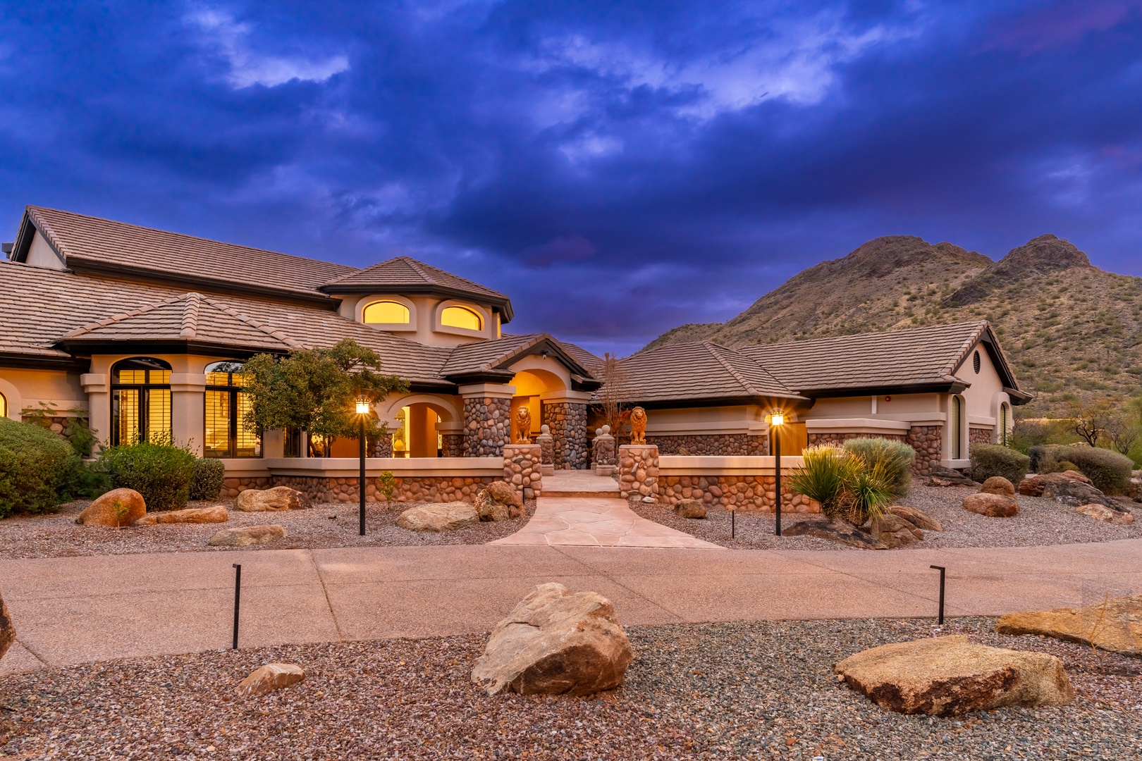 Stunning desert property nestled at the base of rugged mountain peaks, featuring elegant architecture and warm evening lighting that creates an inviting arrival experience.