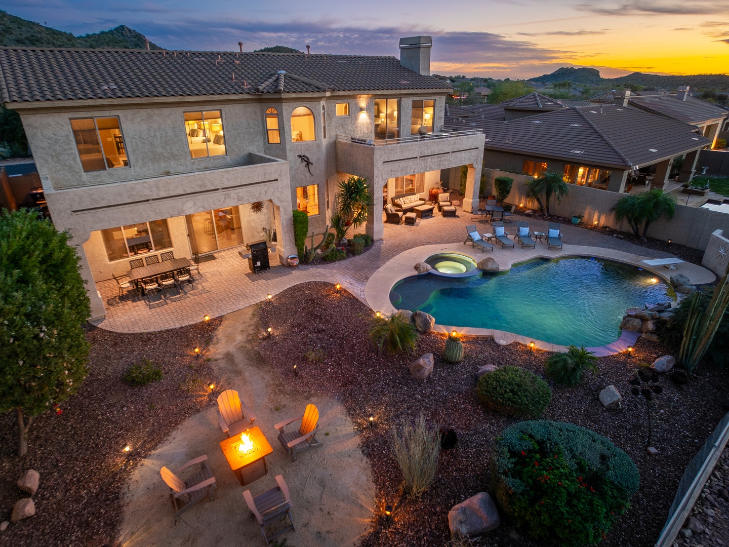 Aerial view of a desert luxury home at sunset, showcasing a sparkling pool, hot tub, fire pit gathering area, and mountain-framed patio under a vibrant evening sky.