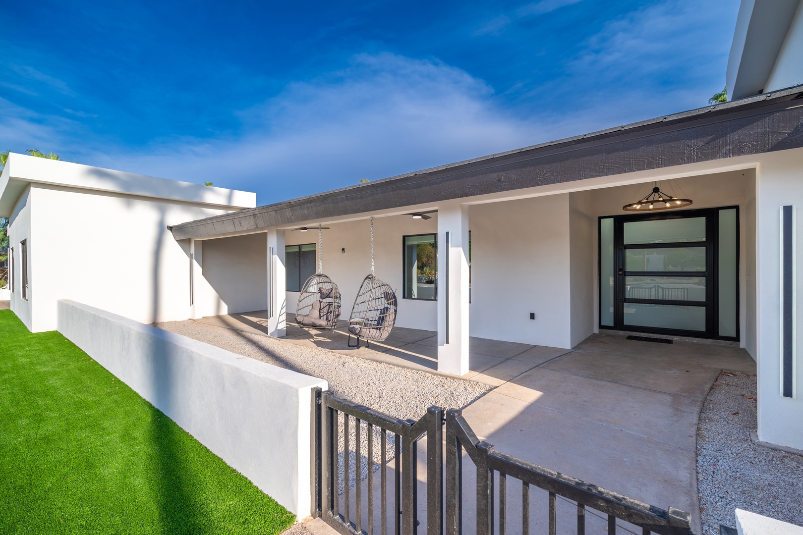 Modern single-level property featuring a covered patio with hanging chairs, clean white exterior, and manicured lawn—an inviting entrance to your desert retreat.