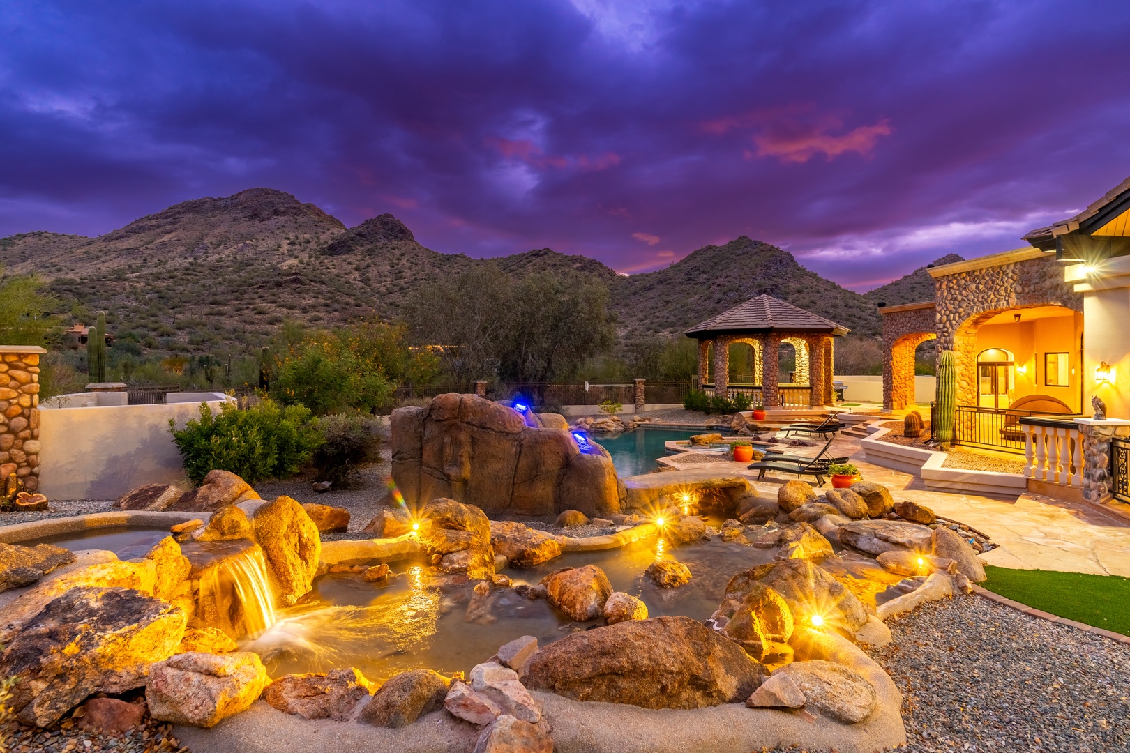 A stunning desert property nestled against mountain peaks, featuring a tranquil water feature with cascading waterfalls and a serene pool, all illuminated under a dramatic twilight sky.