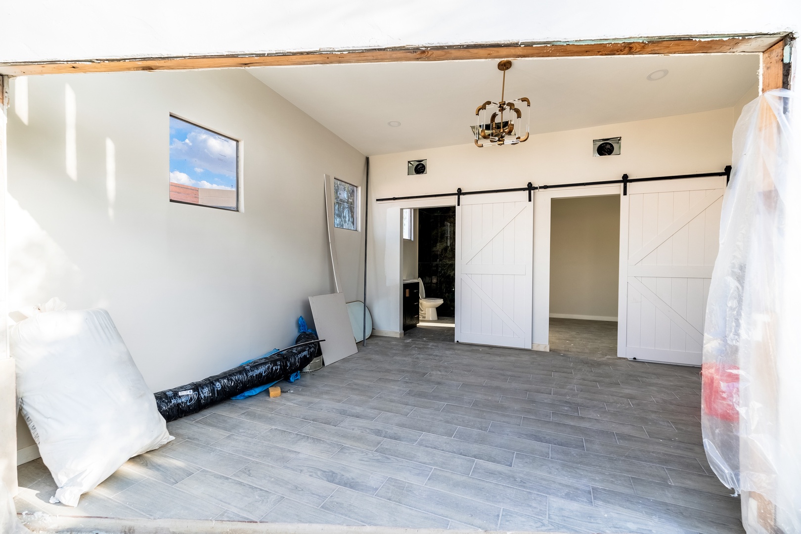 A spacious room under renovation features modern barn doors opening to private areas, exposed beam details adding rustic charm, and clean tile flooring ready to welcome your stay.