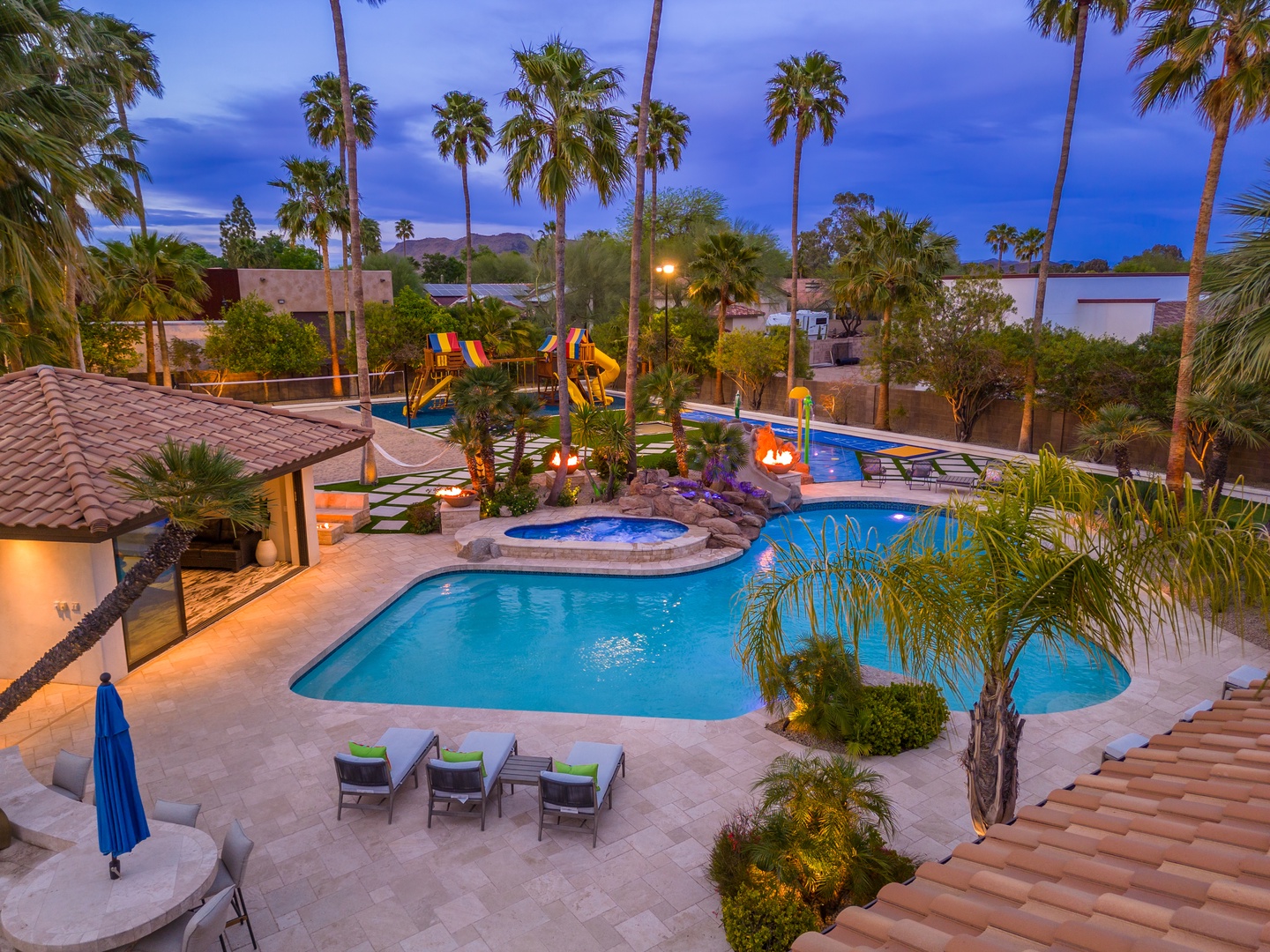 A stunning resort-style backyard retreat featuring multiple pools, water slides, and a spa nestled among towering palms under twilight skies.