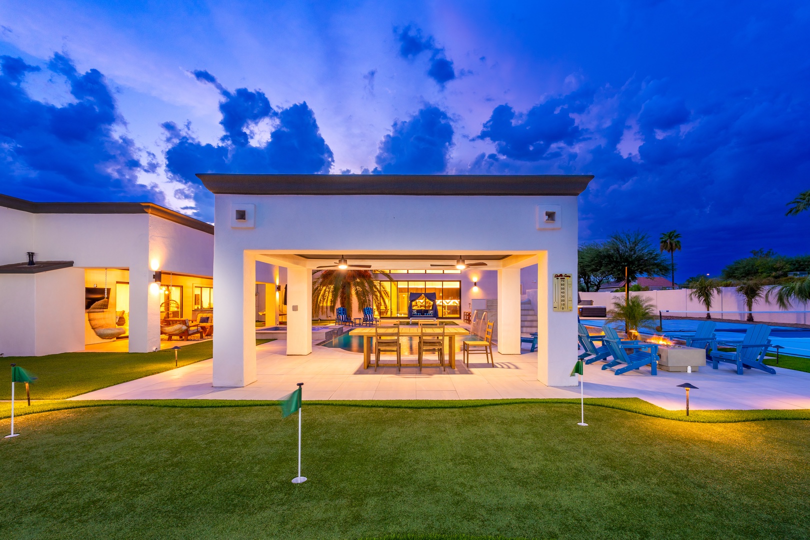 Modern vacation home with covered patio, private pool, and miniature golf course set against a dramatic twilight sky, surrounded by lush tropical landscaping and palm trees.