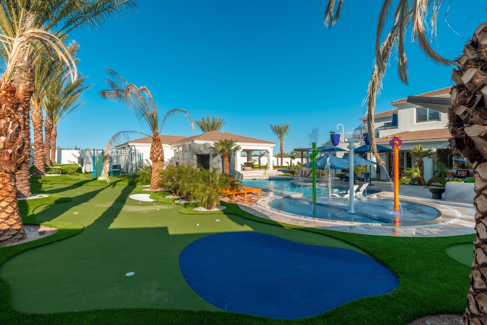 Resort-style backyard with splash zone, mini golf, and lush palms under clear desert skies—perfect for family fun in the sun.