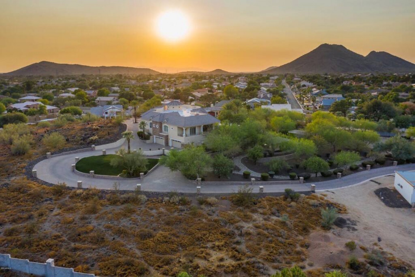 Stunning aerial view captures the property nestled in a peaceful desert neighborhood, surrounded by majestic mountains and golden sunset skies.