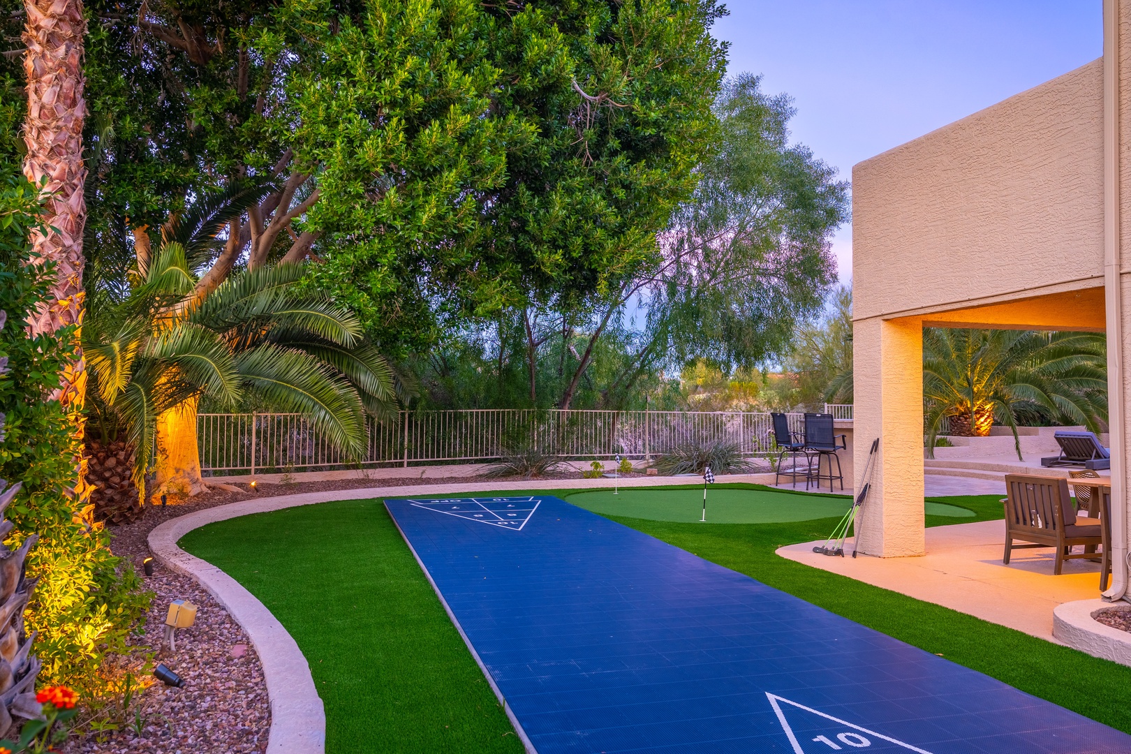 A custom putting green awaits your perfect stroke in this beautifully lit backyard, framed by mature palms and covered patio seating for post-game relaxation.
