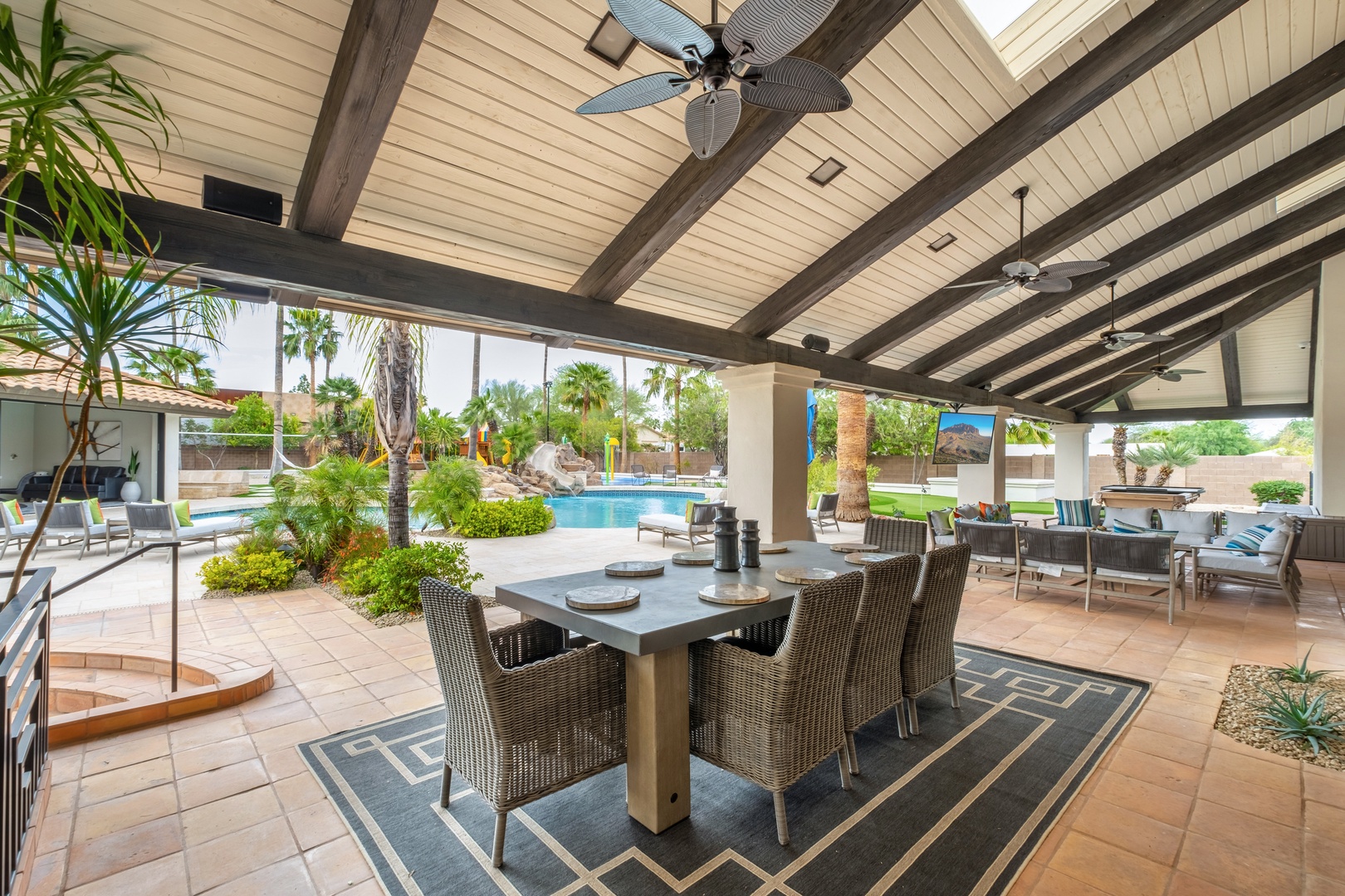 Expansive covered patio with vaulted ceilings, ceiling fans, and elegant wicker dining for eight. Beyond, your sparkling pool awaits, framed by palms and lush desert landscaping—your perfect indoor-outdoor retreat.