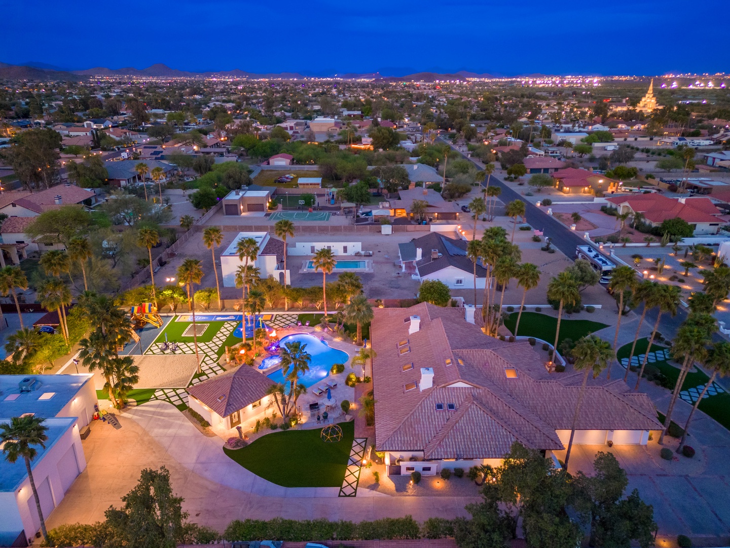 An aerial view of the property at twilight, showcasing a stunning resort-style pool area, lush palm trees, and expansive outdoor spaces nestled in a quiet residential neighborhood.