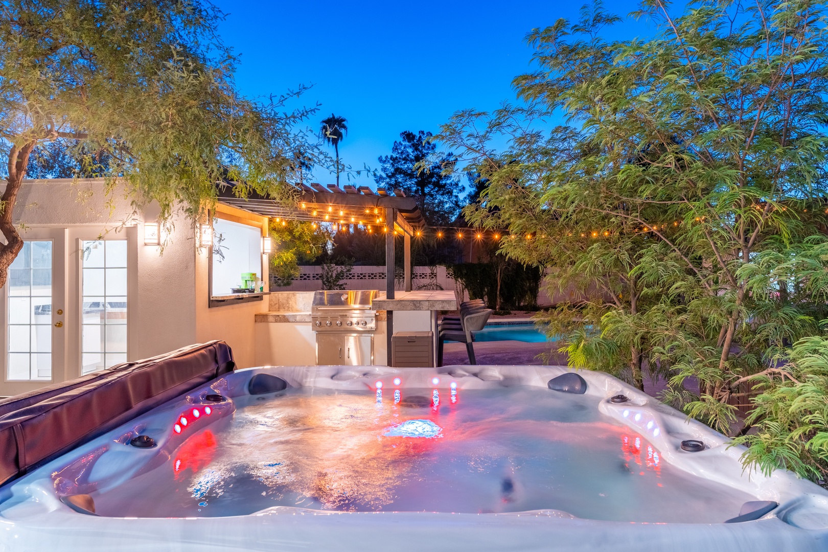Unwind under the stars in your private hot tub, surrounded by lush greenery and ambient lighting. The outdoor kitchen and pool complete your perfect evening retreat.