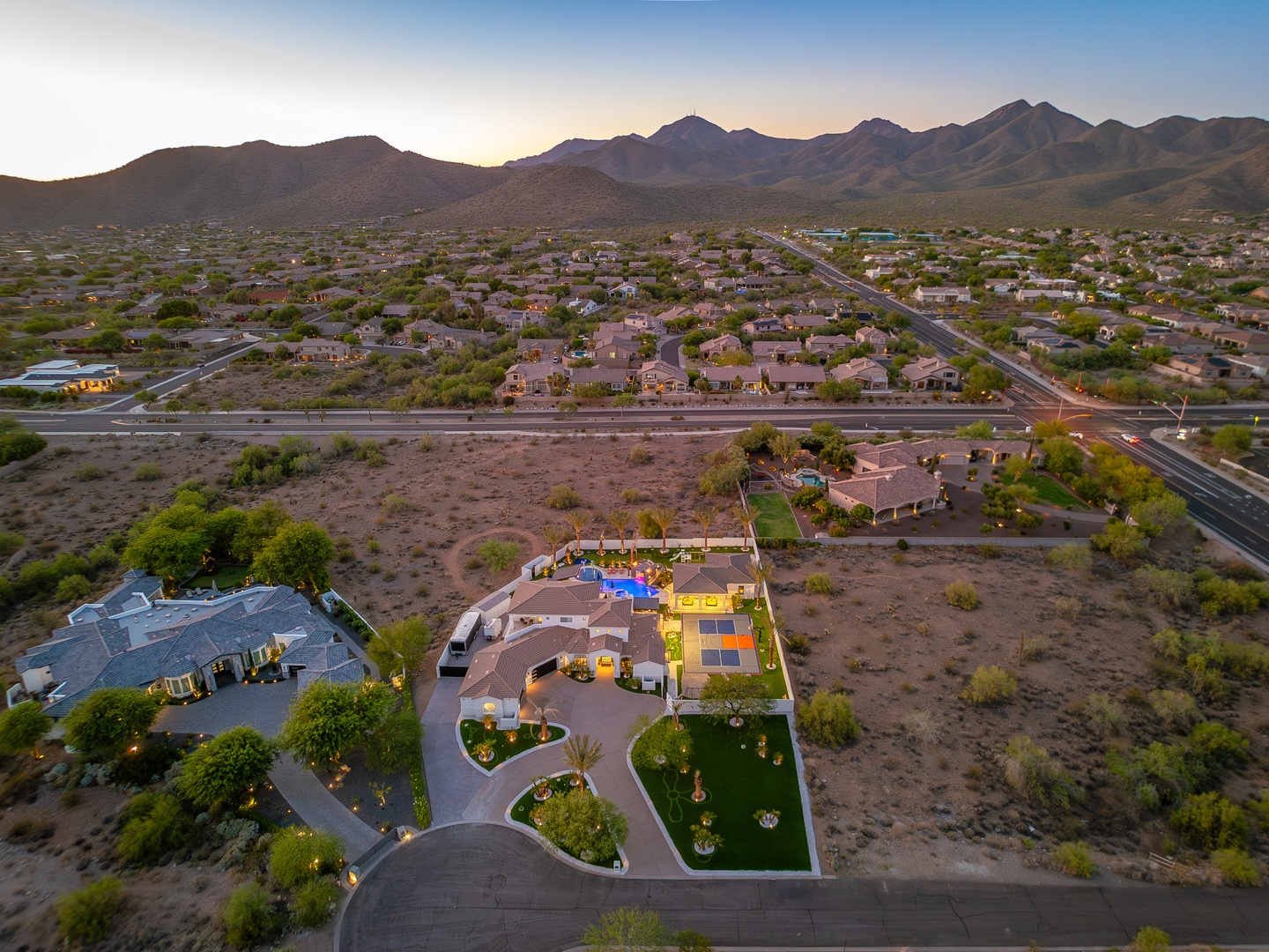 Aerial view of a desert retreat surrounded by mountain ranges, showcasing the property's unique architecture and lush landscaping amid the arid Arizona landscape at dusk.