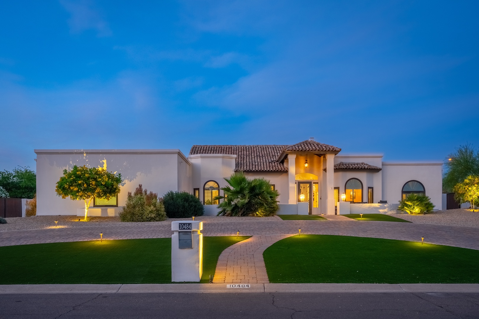 A stunning Spanish-style villa with warm lighting illuminates the entrance at dusk, featuring manicured landscaping and a welcoming brick pathway leading to the main entry.