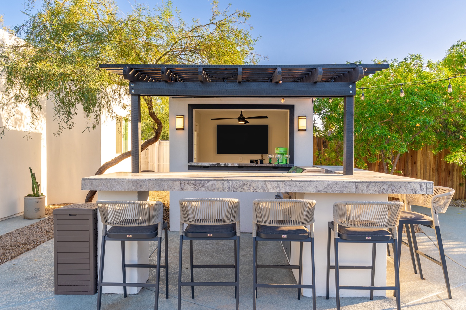 This modern outdoor bar area features a polished concrete counter with stylish seating under a pergola, surrounded by lush greenery—your perfect spot for entertaining guests.