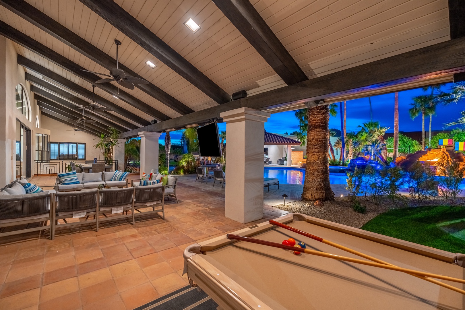 Unwind in your expansive covered patio with pool table, comfortable lounge seating, and stunning twilight views of the sparkling pool and lush tropical landscaping—perfect for entertaining or relaxing.