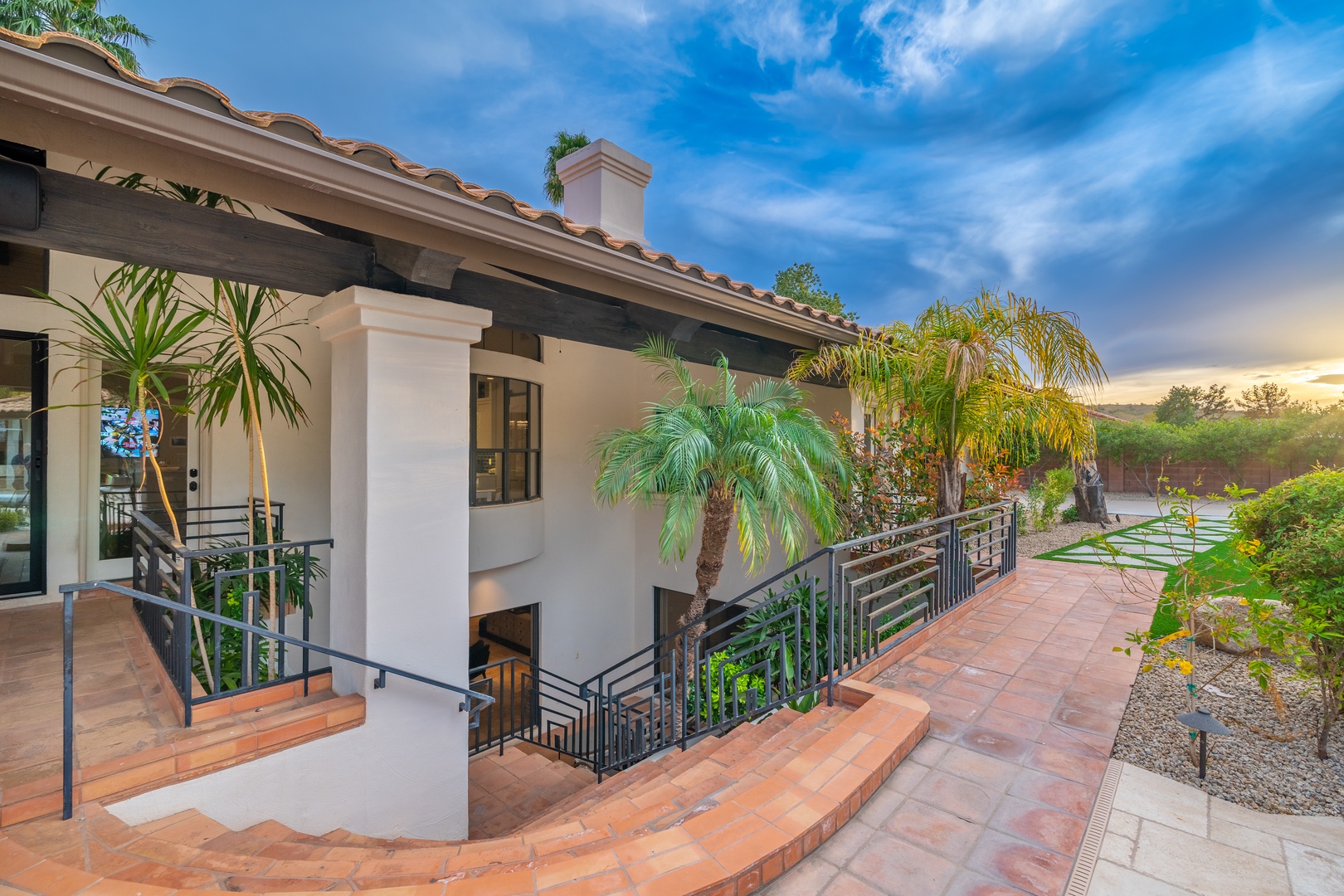Spacious two-story villa with Mediterranean architecture, featuring terracotta tile roofing, lush palm landscaping, and a private terrace overlooking desert scenery. The property is framed by vibrant tropical greenery and a serene natural backdrop.