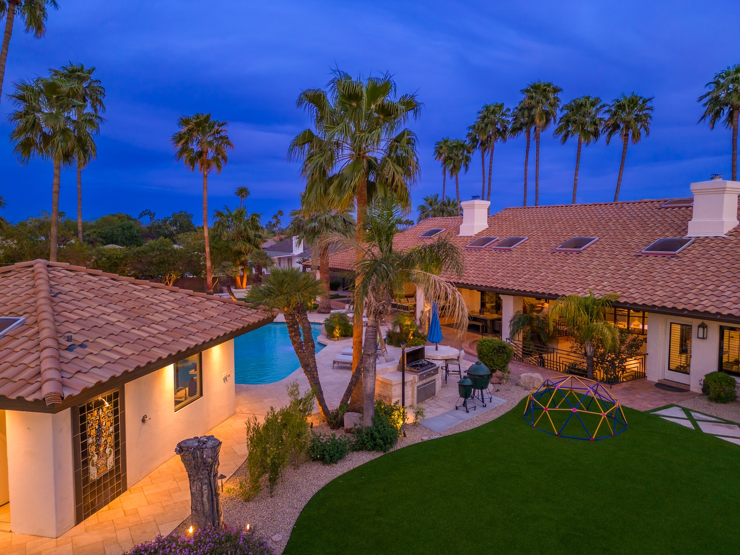 Aerial view of a Spanish-style villa at dusk, featuring a sparkling pool, palm-lined grounds, outdoor BBQ area, and children's playground surrounded by lush landscaping and warm ambient lighting.