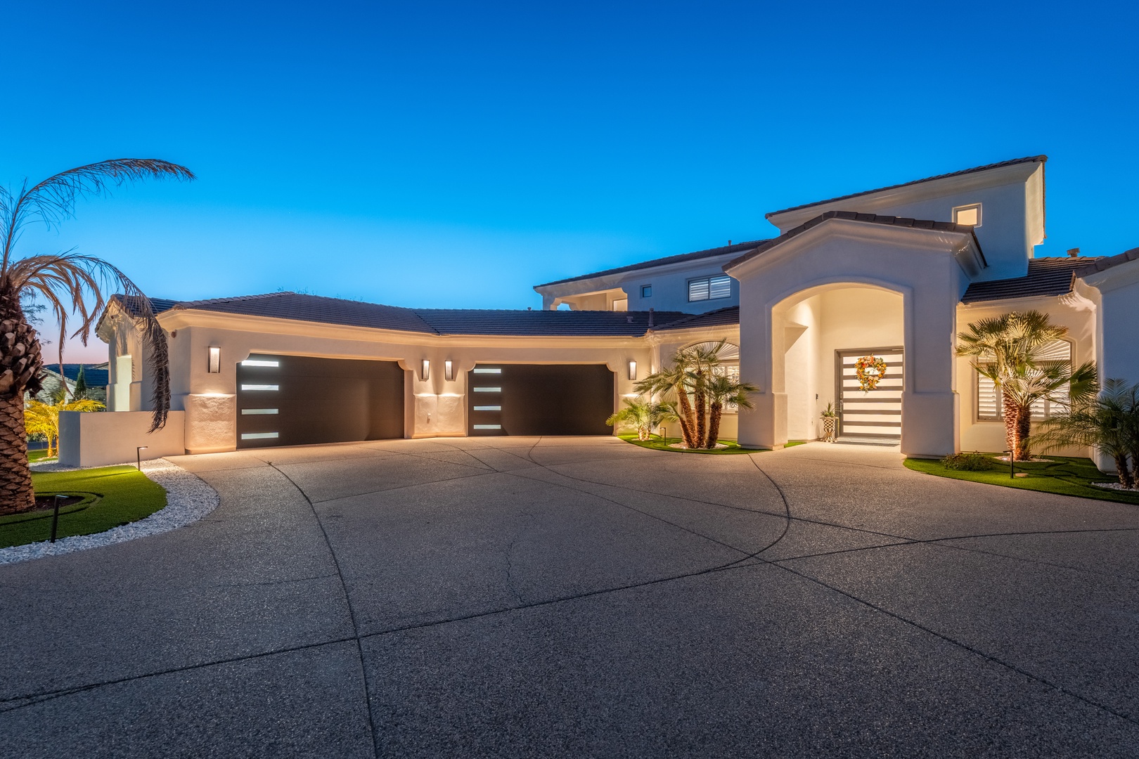 This stunning contemporary property features an elegant arched entrance and modern facade, complemented by stylish landscaping with palm trees and ambient lighting. The spacious driveway provides ample parking.