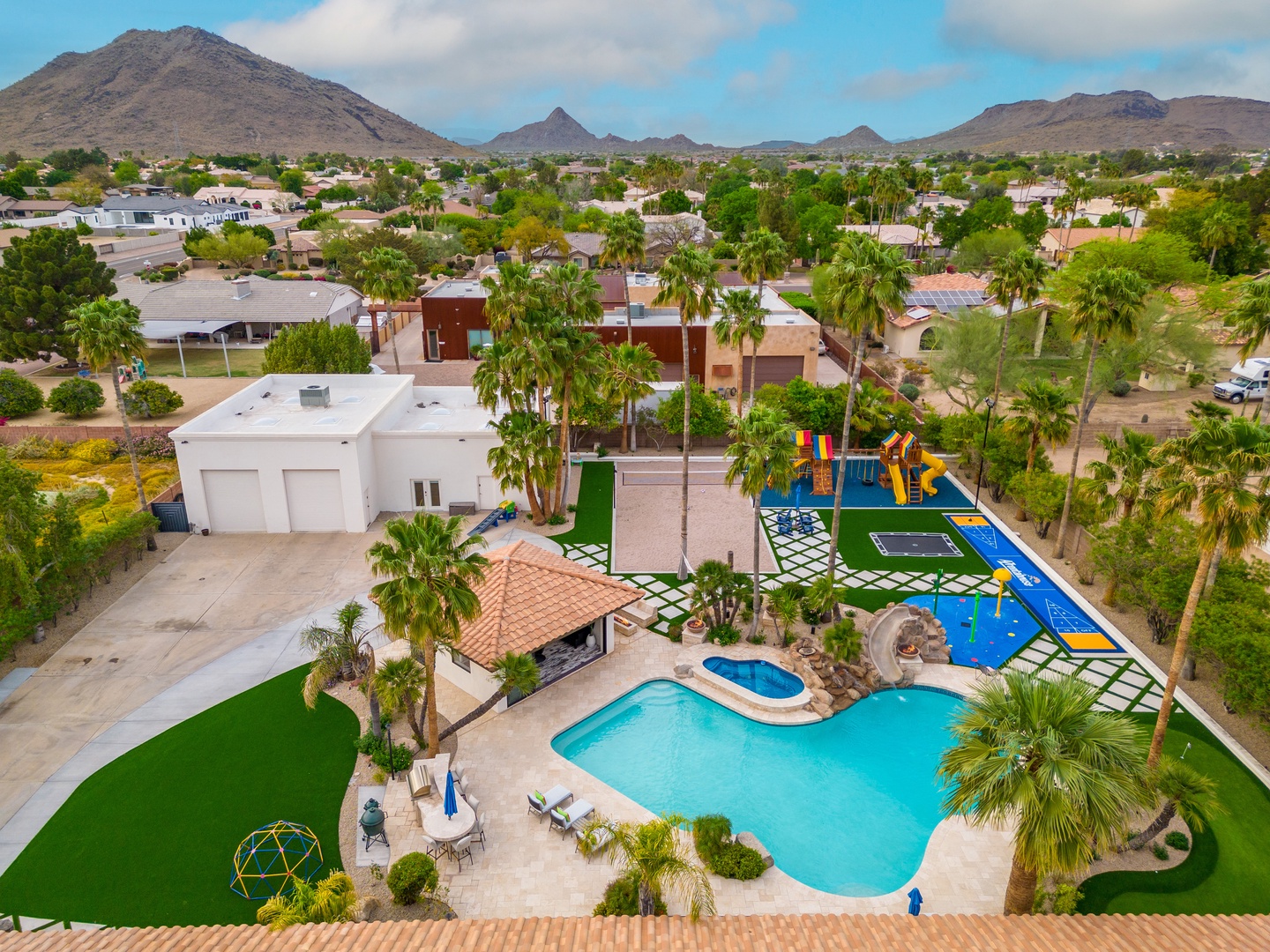 An aerial view of the property showcasing a sparkling pool, vibrant sports courts, playground equipment, and mountain vistas defining the desert residential neighborhood.