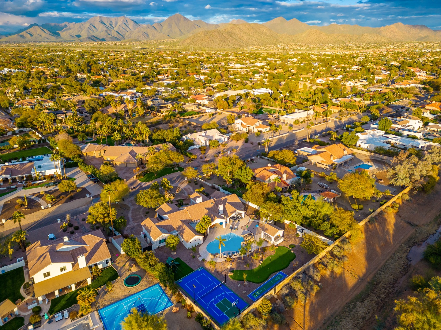 Aerial view captures the property nestled in a sunny desert neighborhood, with majestic mountains framing the horizon and tennis courts nearby.